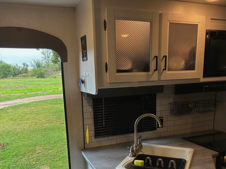 RV rental kitchen: sink, mesh cabinets, microwave, keys on hooks, and a window looking out to a green field.. Keystone RV Hideout 2022