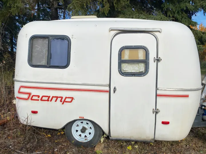 White Scamp camper trailer for rv rental. A compact, rounded travel trailer with windows and a side door, parked outdoors.. Scamp 13' 1987