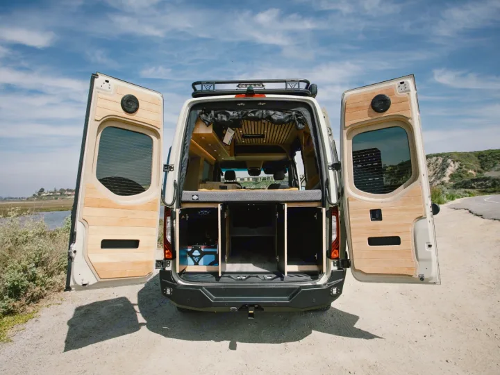 Explore with this RV rental! Rear view of a camper van with open doors, revealing its wood-paneled interior and storage, parked outdoors by water.. Vanspeed Vanspeed Class B 2021