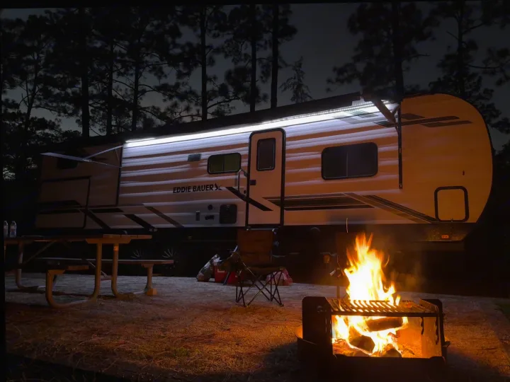 An Eddie Bauer rv rental shines bright at a cozy campsite, with a crackling campfire, picnic table, and chairs under night trees.. Heartland RVs Eddie Bauer 2026