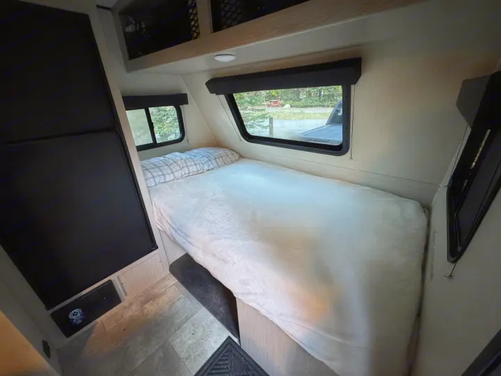 Sleep comfortably in this RV rental! Interior view of a bed with plaid pillows, large windows revealing a peaceful campsite.. Coachmen Apex Remote 16R 2023