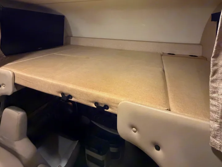 Overhead bunk bed of an RV rental, with a TV on the left, soft cushions, and cup holders. Driver's seat visible below.. Thor Freedom Elite 2017