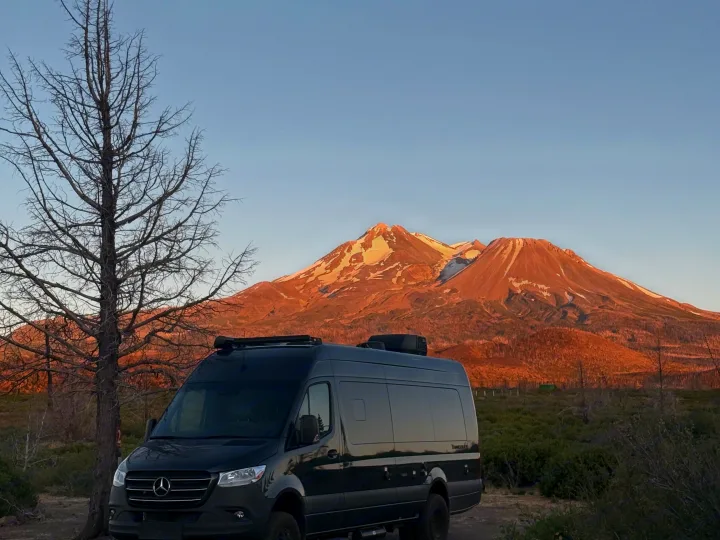 Explore mountains with an RV rental! A black camper van sits before Mount Shasta's golden, snow-dusted peak at sunset.. Thor Tranquility 2025