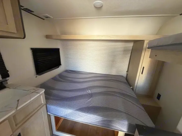 RV rental bedroom: main bed with gray mattress, upper bunk, small sink vanity, and window with dark blind.. Coachmen Shasta 2027
