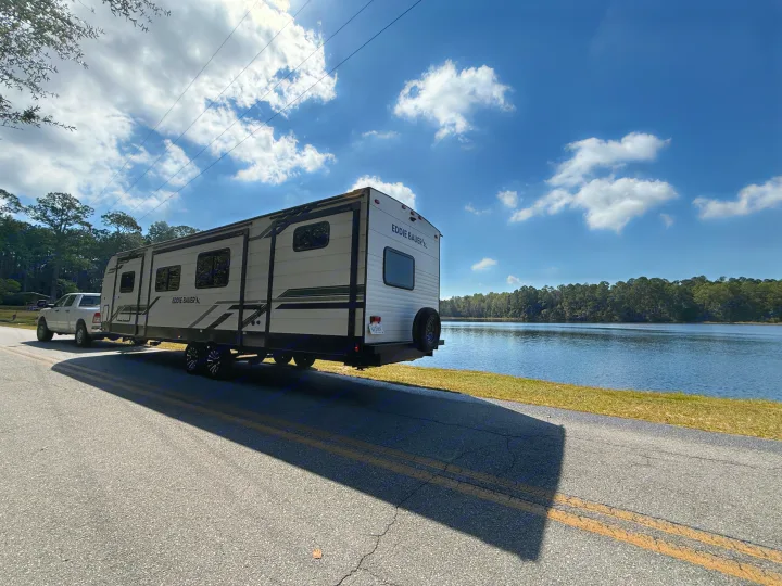 An Eddie Bauer travel trailer (RV rental) and pickup truck are parked on a road beside a serene lake under a sunny sky.. Heartland RVs Eddie Bauer 2026