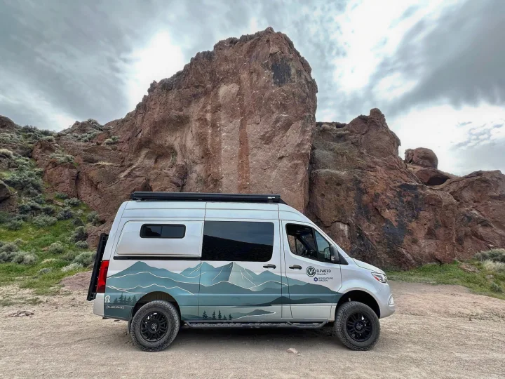 An RV rental camper van, adorned with mountain graphics, is parked on a dirt path amidst majestic rock formations, ideal for adventure.. Mercedes-Benz Sprinter 2023