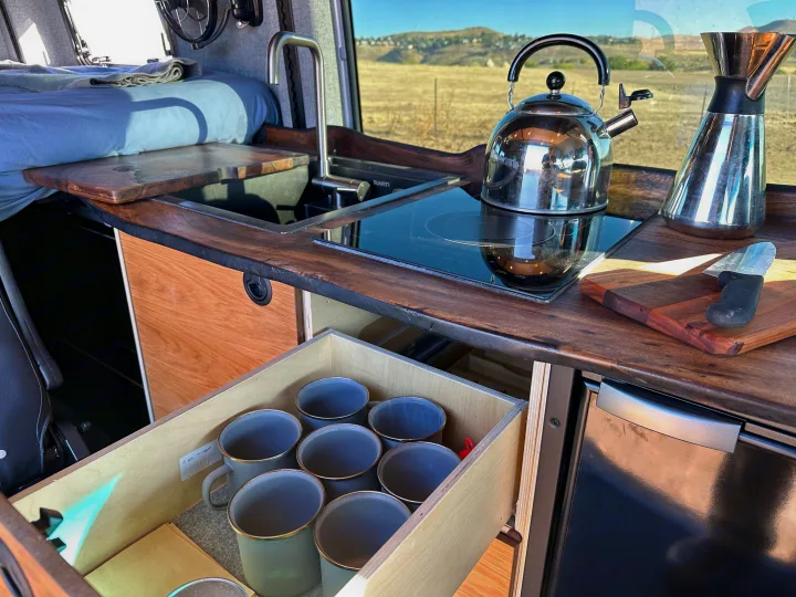 Compact rv rental kitchenette with induction cooktop, sink, and mugs in an open drawer, offering a scenic view.. Mercedes-Benz Sprinter 2023