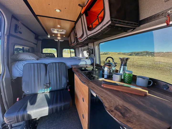 RV rental: Cozy camper van interior showing a full kitchen, comfortable bed, rear seating, & sunny outdoor landscape view.. Mercedes-Benz Sprinter 2023