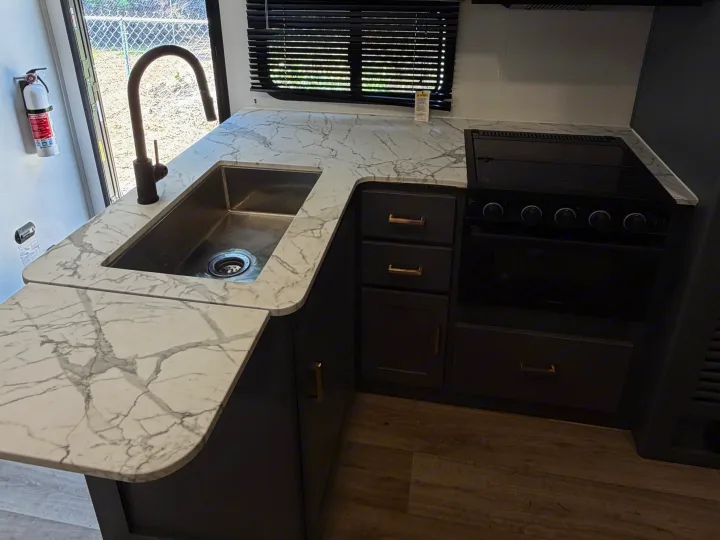 Modern RV rental kitchen features marble-look countertop, stainless steel sink, black stove, dark cabinets, and safety fire extinguisher.. Keystone RV Bullet Crossfire 2024