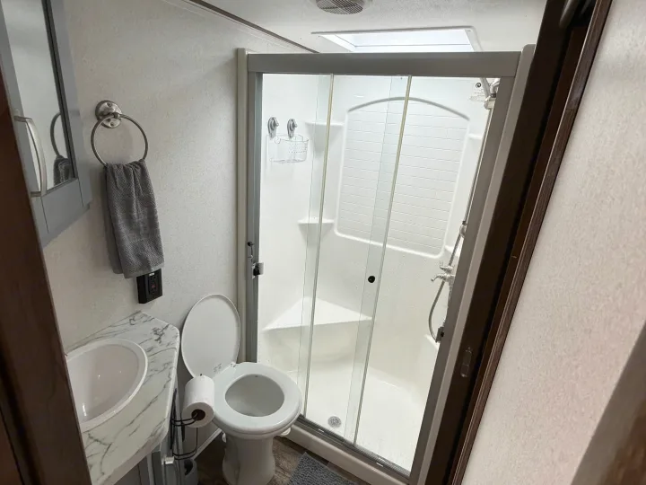 This RV rental bathroom features a compact toilet, sink with marble-patterned counter, and a glass-enclosed shower with a skylight.. Keystone Fuzion 2020
