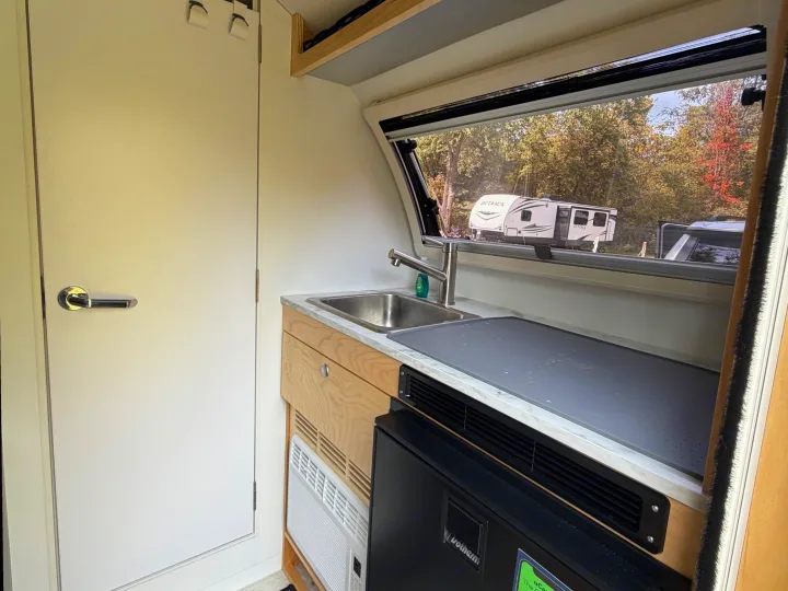 Compact RV rental kitchen interior with sink, counter, fridge, and AC. Another RV is visible through the window.. nuCamp T@B 2026