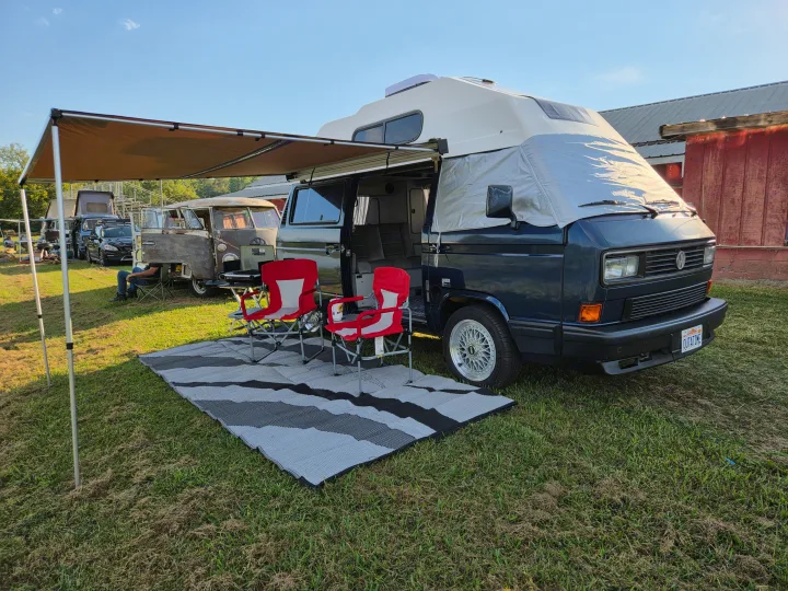 A blue VW camper van, set up for outdoor relaxation with an awning, chairs, and rug on grass. Great for a scenic rv rental trip.. Volkswagen Vanagon 1990