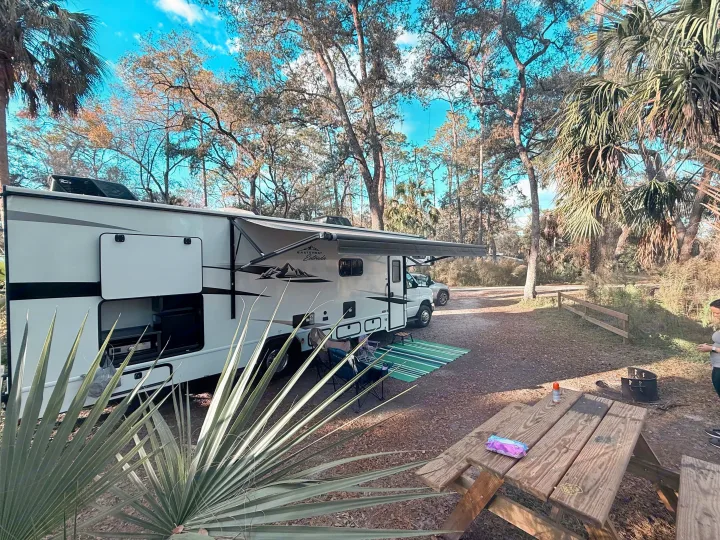 White East to West Entrada RV rental, with an outdoor kitchen & awning, parked at a shaded campsite with a picnic table.. East to West Entrada 2025