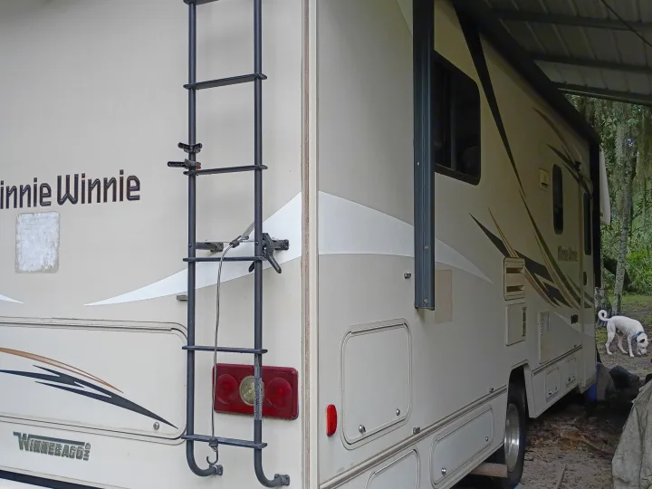 Winnebago Minnie Winnie RV rental in a wooded setting. Features a rear ladder, side awning, and a white dog nearby.. Winnebago Minnie Winnie 2016