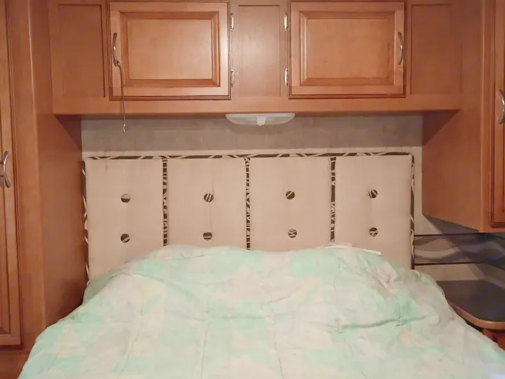 Cozy RV rental bedroom featuring overhead wooden cabinets, a tufted headboard, and a light green patterned blanket.. Winnebago Minnie Winnie 2016
