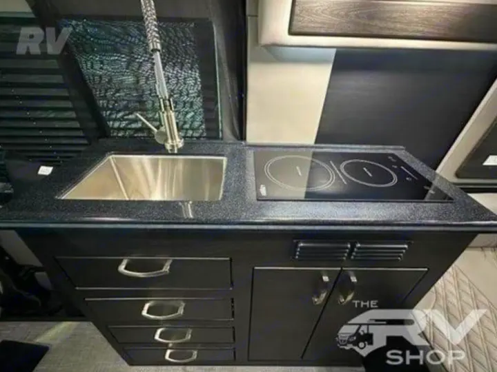 RV rental kitchen: stainless steel sink, black induction stovetop, and dark cabinets. A compact, modern cooking area for your adventures.. Midwest Automotive Designs Passage 2024