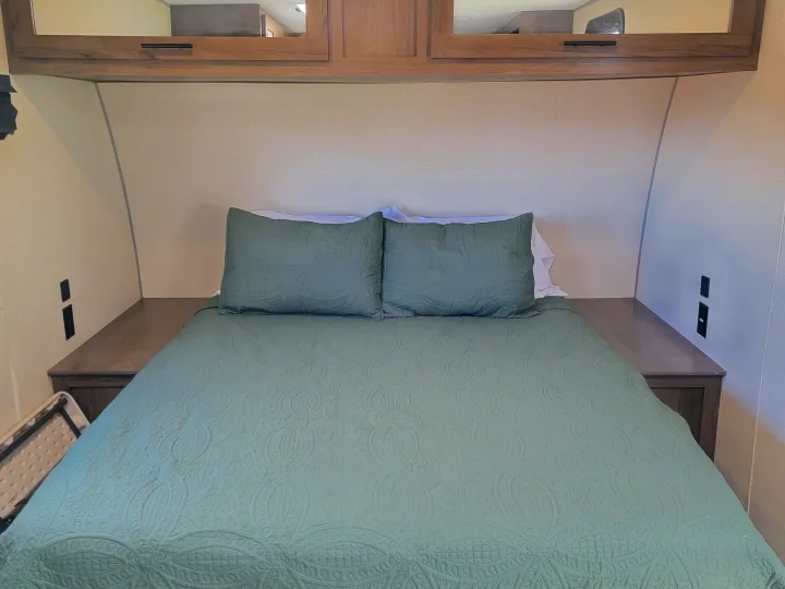 Cozy RV rental bedroom features a green quilted bed, built-in nightstands with outlets, and overhead cabinets.. Forest River Avenger 2020