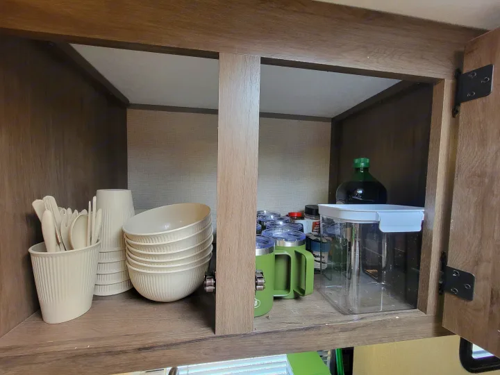 Inside an RV rental cabinet: organized cream-colored dishes, utensils, green mugs, and pantry items, ideal for compact travel storage.. Forest River Avenger 2020