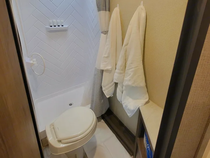 Compact RV rental bathroom features a white herringbone shower, toilet, and hanging towels, with mini toiletries.. Forest River Avenger 2020