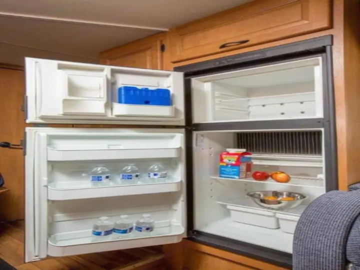 Open RV rental refrigerator and freezer, stocked with milk, fruit, water, and an ice pack in a wooden interior.. Thor Majestic 2023