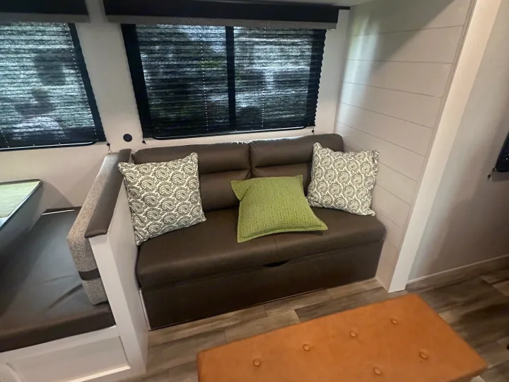 Modern RV rental living space with a brown sofa, accent pillows, windows with blinds, and a dining booth on the left.. Colt Open range 26dbs 2026
