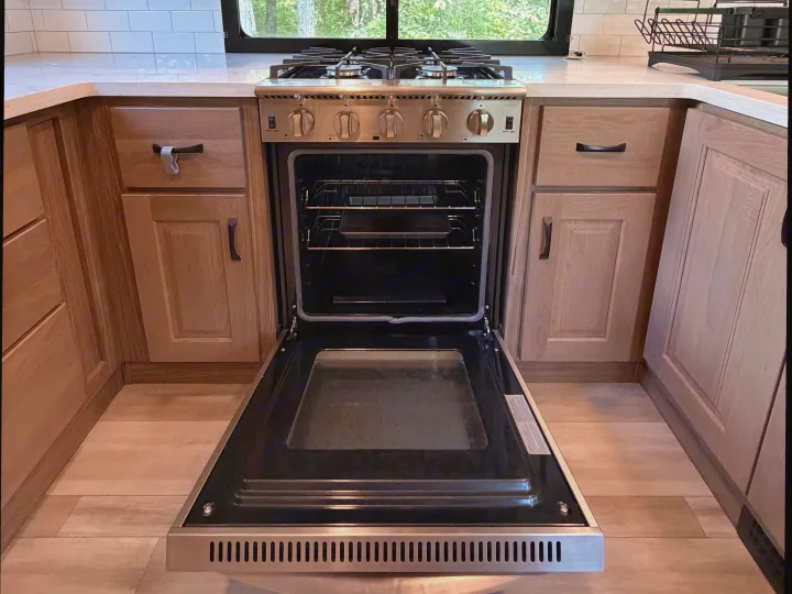 Inside a modern **rv rental** kitchen, an open oven with a gas stove sits between light wood cabinets and a subway tile backsplash.. Alliance RV Paradigm 2024
