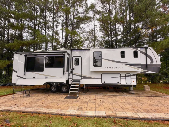 This Paradigm fifth-wheel RV rental is set up on a paved patio, with steps leading to the door. Pine trees line the background.. Alliance RV Paradigm 2024