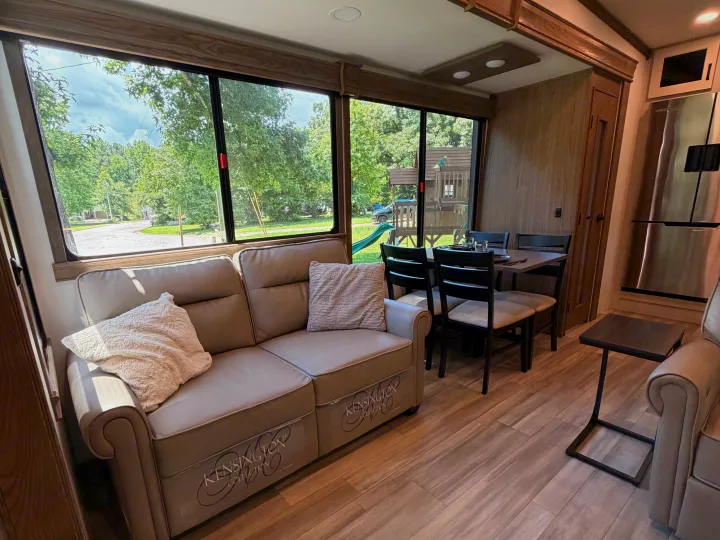 Spacious RV rental interior featuring a comfy sofa, dining table, kitchen, and large windows revealing a lush outdoor view.. Alliance RV Paradigm 2024