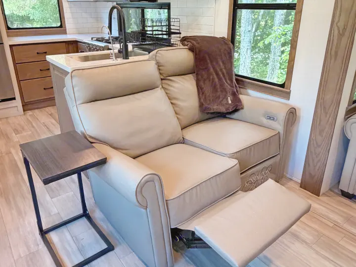 Cozy rv rental interior featuring a light-colored reclining loveseat, a small side table, and a kitchenette, with trees visible outside the window.. Alliance RV Paradigm 2024