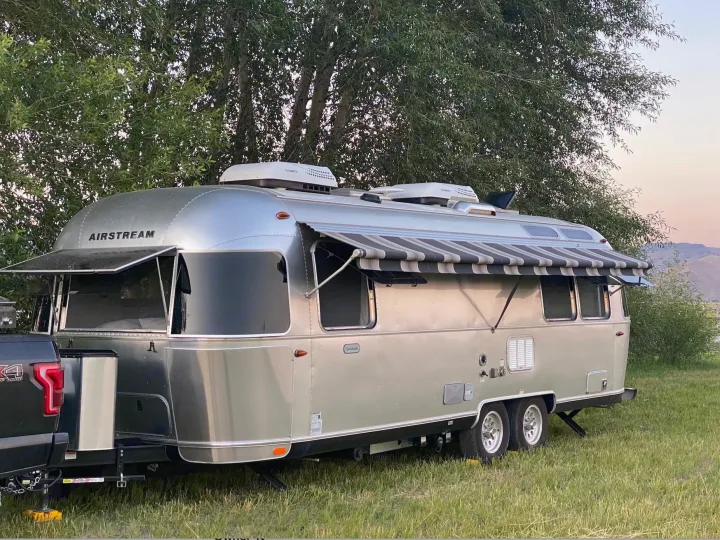 This silver Airstream travel trailer (rv rental) is parked in a grassy field with a truck, trees, & mountains.. Airstream Globetrotter 2019