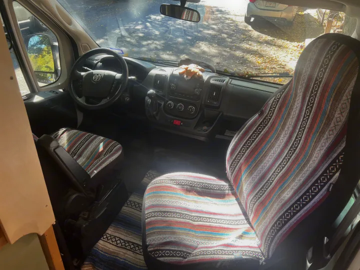 Inside an RV rental van, colorful patterned seat covers and dashboard controls are ready for a journey. View through windshield.. Dodge Promaster 2500 high roof 2019