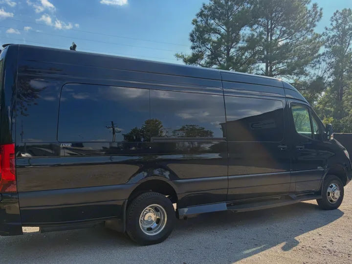 Black Mercedes-Benz Sprinter LUXE van, seen from the side with dark tinted windows reflecting sky & trees, ideal for RV rental.. Midwest Automotive Designs Luxe Cruiser 2025
