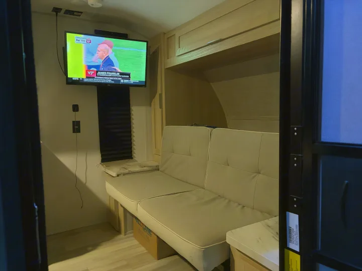 Cozy RV rental interior featuring a TV showing a college football game (VT), a beige futon, and built-in storage.. Forest River Wildwood FSX Platinum 2023