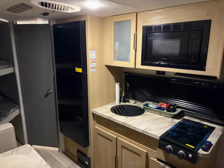 Modern rv rental interior: kitchen area with microwave, refrigerator, stovetop, sink, and adjacent bunk beds.. Forest River Wildwood FSX Platinum 2023