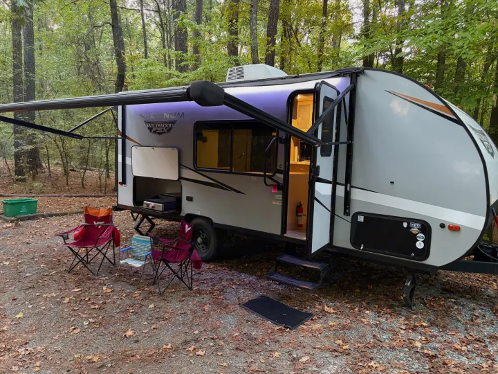 Enjoy an RV rental camping trip! This travel trailer features an extended awning, outdoor kitchen, and chairs at a wooded campsite.. Forest River Wildwood FSX Platinum 2023