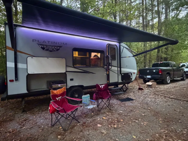 Cozy evening at a wooded campsite with an RV rental, featuring an illuminated travel trailer, camping chairs, and a pickup truck.. Forest River Wildwood FSX Platinum 2023