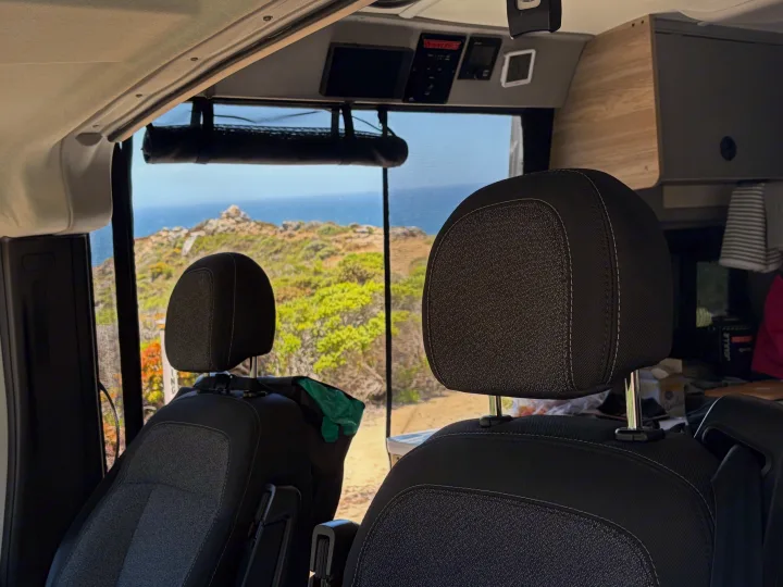Interior of an RV rental, featuring front seats, dashboard controls, and a stunning ocean coastline view outside.. Winnebago Solis 2025