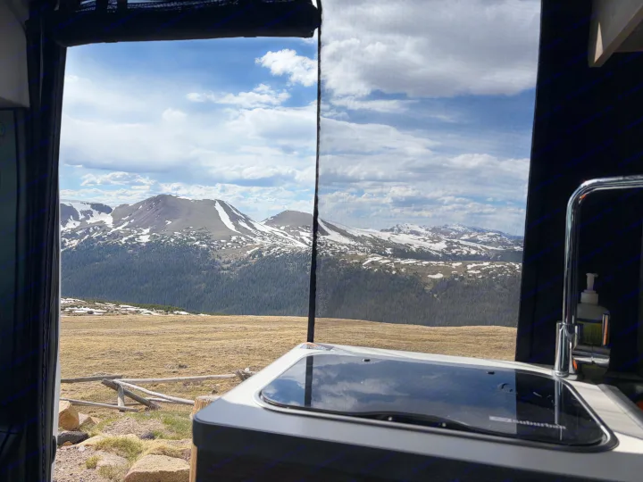 Panoramic mountain view from an RV rental campervan. Features an interior kitchen and forested, snow-capped peaks.. Winnebago Solis 2025