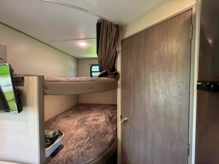 This RV rental features bunk beds with gray bedding, a small window with curtains, and a door, all in neutral tones.. Coachmen Catalina Summit 261BH 2025