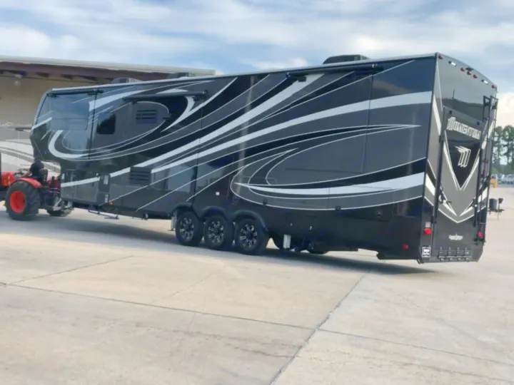 Large black Momentum fifth-wheel RV rental, stylish with white graphics, being moved by a red tractor on concrete.. Grand Design Momentum Toy Hauler 2020