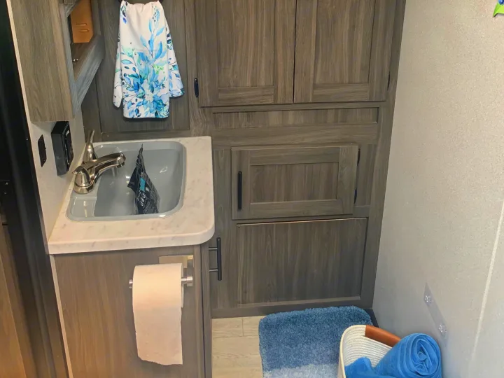 RV rental bathroom with a sink, mirror, wood-look cabinets, TP holder, and blue towels.. Forest River Shasta 2021