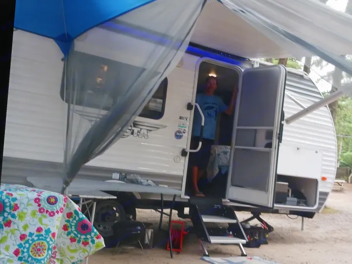 Man in an RV rental with blue awning extended at a dirt and gravel campsite, featuring a vibrant picnic table.. Forest River Shasta 2021