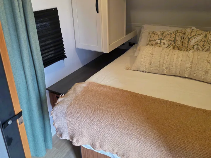 Cozy RV rental bedroom: bed with decorative pillows, textured throw, overhead cabinet, and window with pleated blind/curtain.. Keystone RV Passport Classic 2025