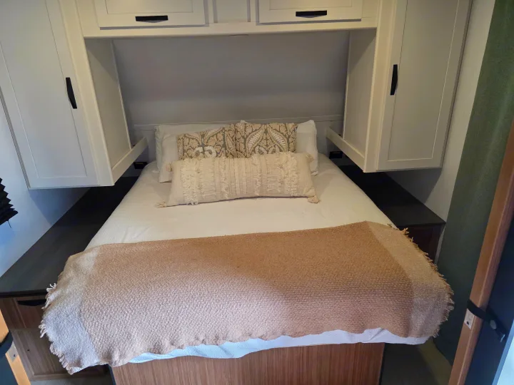 Cozy RV rental bedroom with a white bed, patterned pillows, a throw blanket, and overhead storage cabinets.. Keystone RV Passport Classic 2025