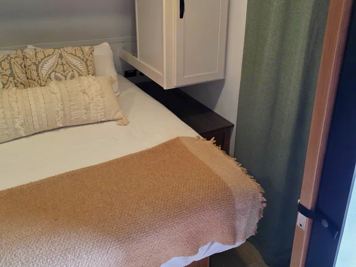 Experience comfort in this RV rental bedroom: a cozy bed with decorative pillows, overhead cabinet, bedside shelf, and green curtain.. Keystone RV Passport Classic 2025