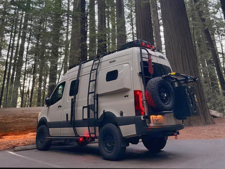 Custom RV rental camper van parked in a majestic redwood forest. Equipped with ladder, roof rack, and rear gear.. Winnebago Revel 2022