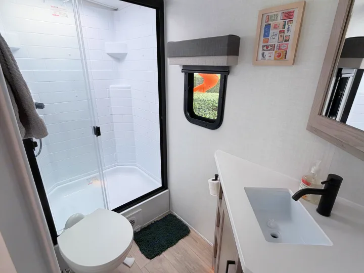 Compact RV rental bathroom with a modern shower, toilet, and vanity sink. A small window offers a glimpse of an outdoor slide.. Forest River 2706BH 2026