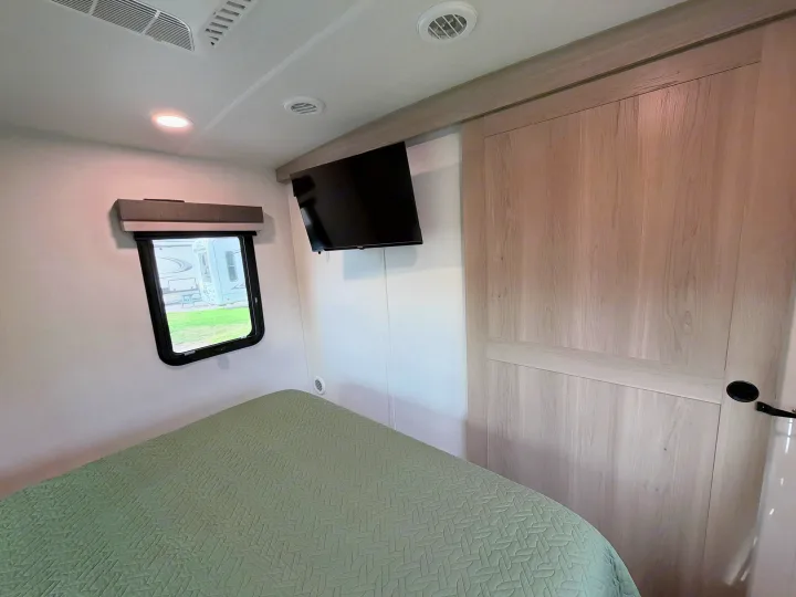 Bright RV rental bedroom: bed with green quilt, window, wall-mounted TV, and light wood closet door.. Forest River 2706BH 2026