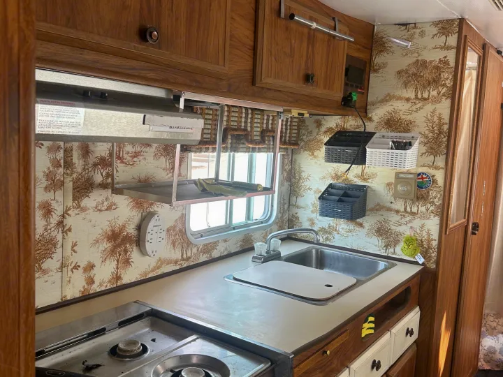 Inside an rv rental, a compact kitchen features wooden cabinets, scenic wallpaper, a sink, and a stovetop. A window is above the counter.. Winnebago Winnebago 1980