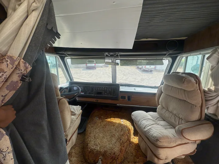 Inside a classic RV rental cockpit, featuring a vintage dashboard, steering wheel, worn seats, and shaggy carpet.. Winnebago Winnebago 1980